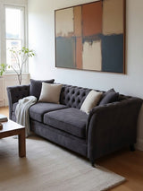 Three-seat grey velvet sofa with tufted backrest and armrests, adorned with two decorative pillows, placed in a bright room featuring a large abstract wall art piece and a wooden coffee table on a light rug