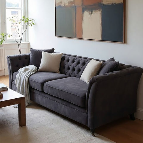 Three-seat grey velvet sofa with tufted backrest, two decorative pillows, and a cozy throw draped over one arm, positioned in a bright room with a large window and a piece of abstract wall art. A wooden coffee table is visible in the foreground