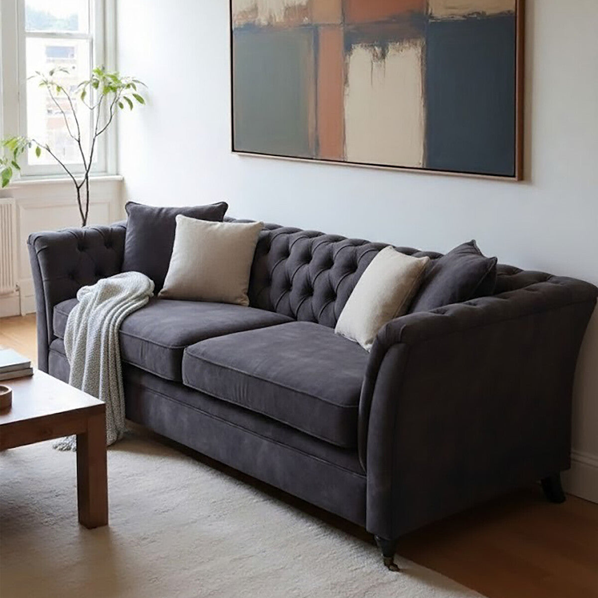 Three-seat grey velvet sofa with tufted backrest, two decorative pillows, and a cozy throw draped over one arm, positioned in a bright room with a large window and a piece of abstract wall art. A wooden coffee table is visible in the foreground