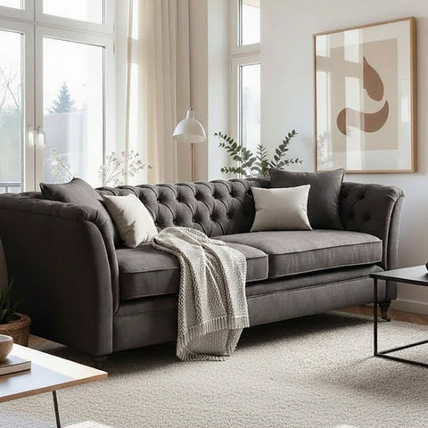 Three-seat grey velvet sofa with tufted backrest, adorned with decorative pillows and a cozy throw blanket, positioned in a bright living room with large windows and modern artwork on the wall. A coffee table sits in front of the sofa on a light-colored rug