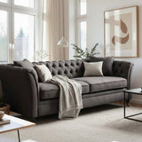 Three-seat grey velvet sofa with tufted backrest, adorned with decorative pillows and a cozy throw blanket, positioned in a bright living room with large windows and modern artwork on the wall. A coffee table sits in front of the sofa on a light-colored rug