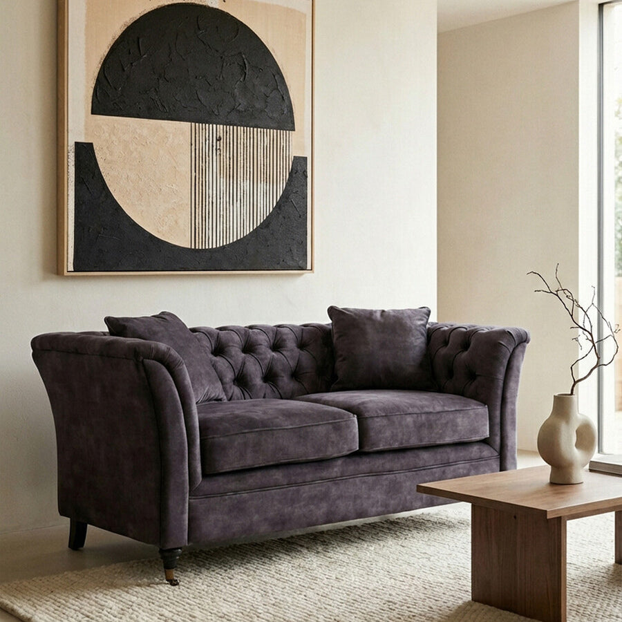 Two-seat grey velvet sofa with tufted backrest and armrests, positioned in a modern living space. A round coffee table made of wood sits in front of the sofa, and a textured area rug is beneath it. A large abstract wall art piece featuring geometric shapes in neutral tones hangs above the sofa, complementing the overall decor. Natural light filters in from a nearby window, enhancing the cozy atmosphere