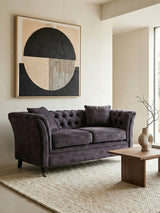 Two-seat grey velvet sofa with tufted backrest and armrests, positioned in a modern living space. A round coffee table made of wood sits in front of the sofa, and a textured area rug is beneath it. A large abstract wall art piece featuring geometric shapes in neutral tones hangs above the sofa, complementing the overall decor. Natural light filters in from a nearby window, enhancing the cozy atmosphere