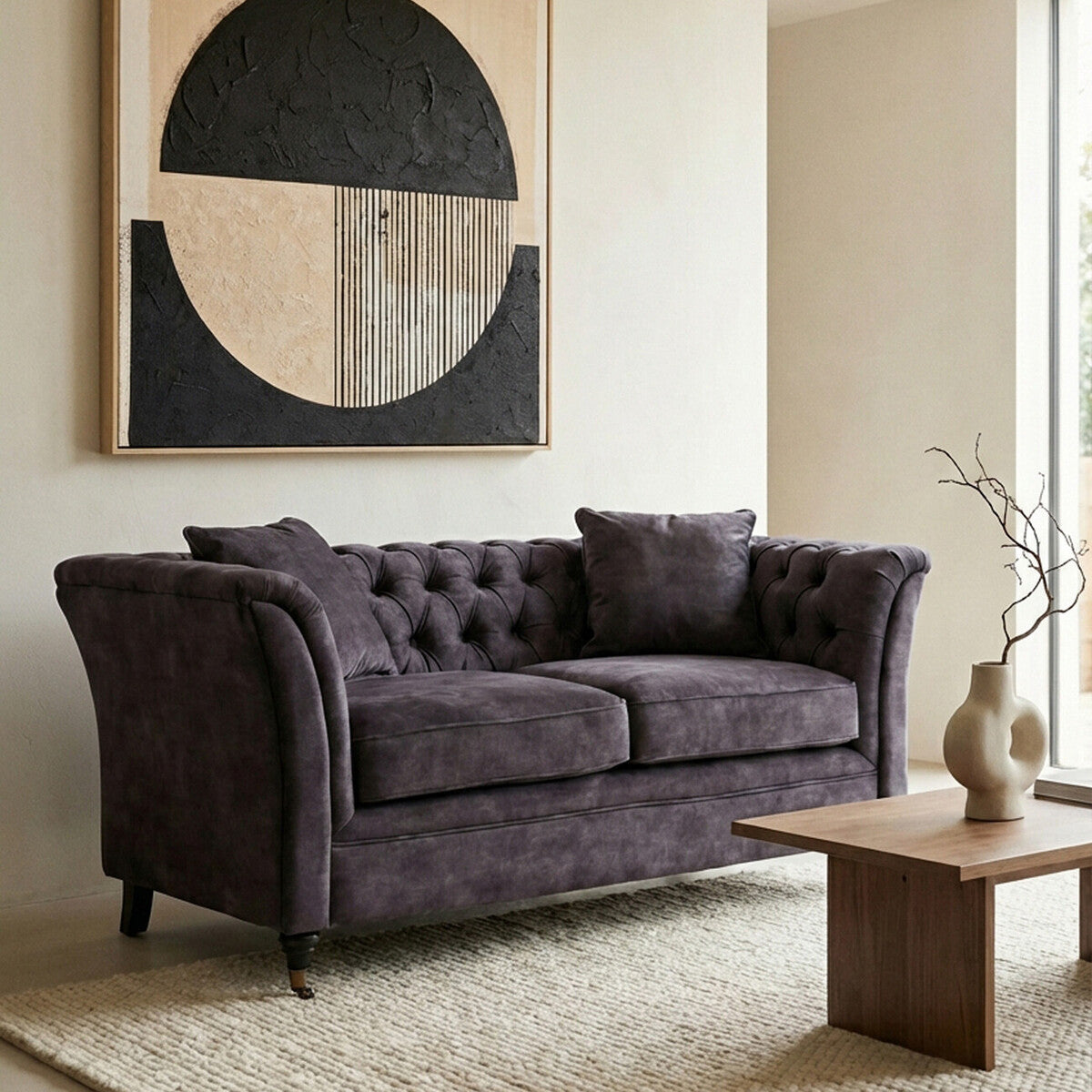 Two-seat grey velvet sofa with tufted backrest and armrests, accompanied by a decorative cushion, placed in a modern living room setting with a geometric wall art piece and a wooden coffee table on a textured rug
