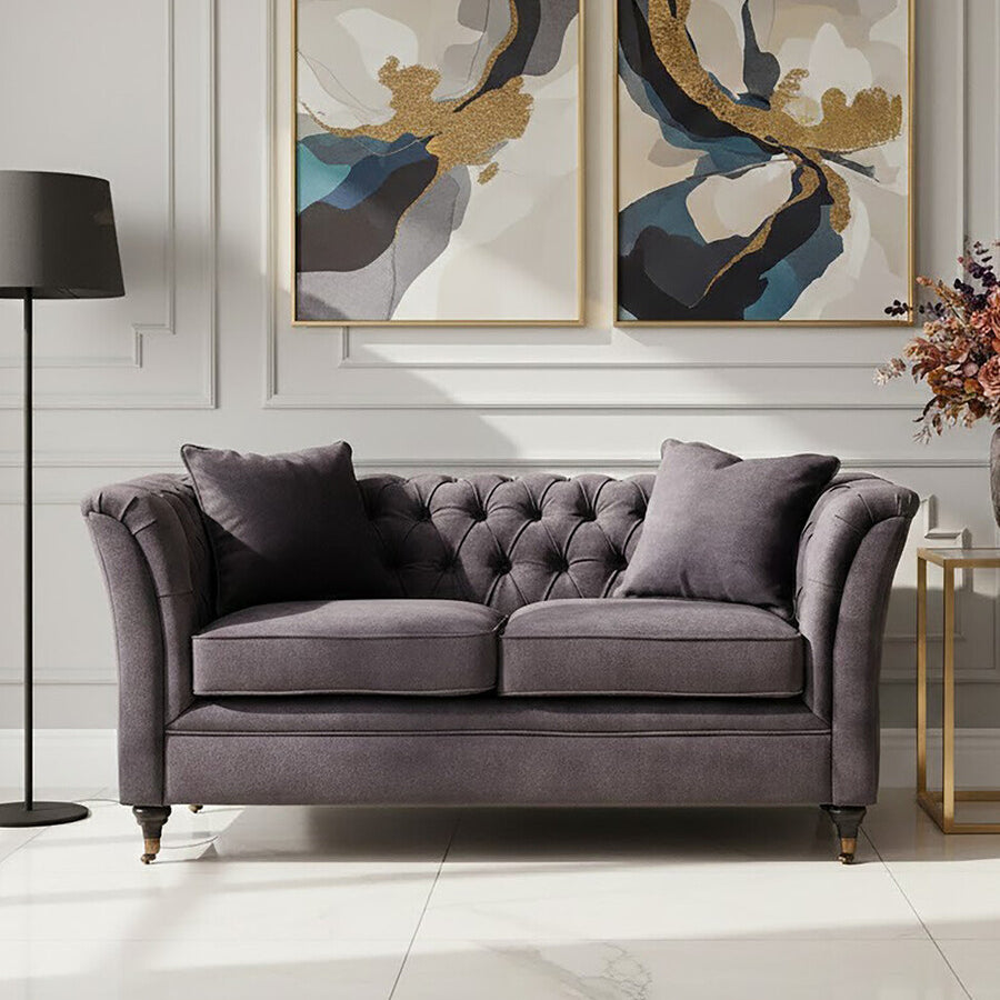 Two-seat grey velvet sofa with tufted backrest and plush cushions, positioned in a stylish living room setting. The sofa is complemented by two decorative pillows and is placed against a wall featuring two abstract art pieces. A modern floor lamp stands nearby, and a small side table holds a vase with flowers. The flooring is light-colored, enhancing the overall elegance of the space