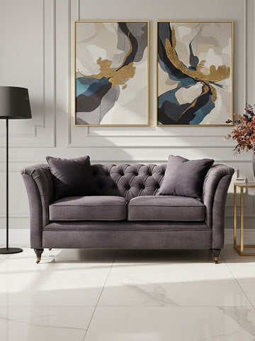 Two-seat grey velvet sofa with tufted backrest and plush cushions, positioned in a stylish living room setting. The sofa is complemented by two decorative pillows and is placed against a wall featuring two abstract art pieces. A modern floor lamp stands nearby, and a small side table holds a vase with flowers. The flooring is light-colored, enhancing the overall elegance of the space