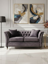 Two-seat grey velvet sofa with tufted backrest and plush cushions, positioned in a stylish living room setting. The sofa is complemented by two decorative pillows and is placed against a wall featuring two abstract art pieces. A modern floor lamp stands nearby, and a small side table holds a vase with flowers. The flooring is light-colored, enhancing the overall elegance of the space