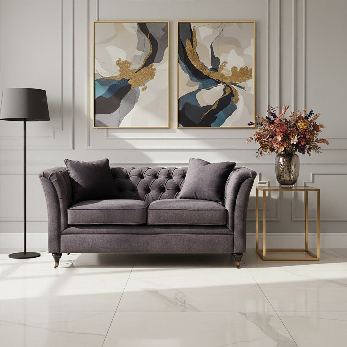 Grey velvet two-seat sofa with tufted backrest, paired with two decorative pillows. A modern floor lamp stands to the left, and a side table with a vase of flowers is positioned on the right. Abstract artwork hangs on the wall behind the sofa, and the floor features light-colored tiles