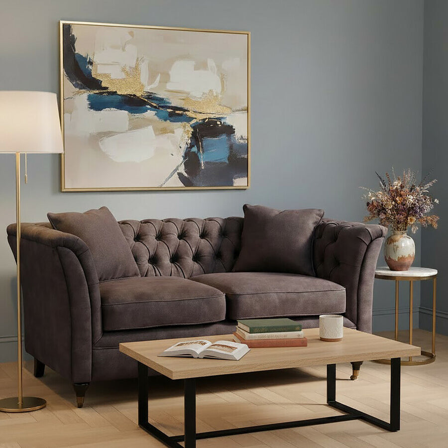 Two-seat grey velvet sofa with tufted backrest and plush cushions, positioned in a cozy living room setting. A wooden coffee table with a black metal frame sits in front of the sofa, and a floor lamp provides warm lighting. A decorative painting hangs on the wall, and a vase with dried flowers is placed on the table. The room features light-colored flooring and a soft color palette