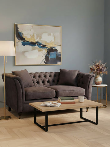 Two-seat grey velvet sofa with tufted backrest and plush cushions, positioned in a cozy living room setting. A wooden coffee table with a black metal frame sits in front of the sofa, and a floor lamp provides warm lighting. A decorative painting hangs on the wall, and a vase with dried flowers is placed on the table. The room features light-colored flooring and a soft color palette