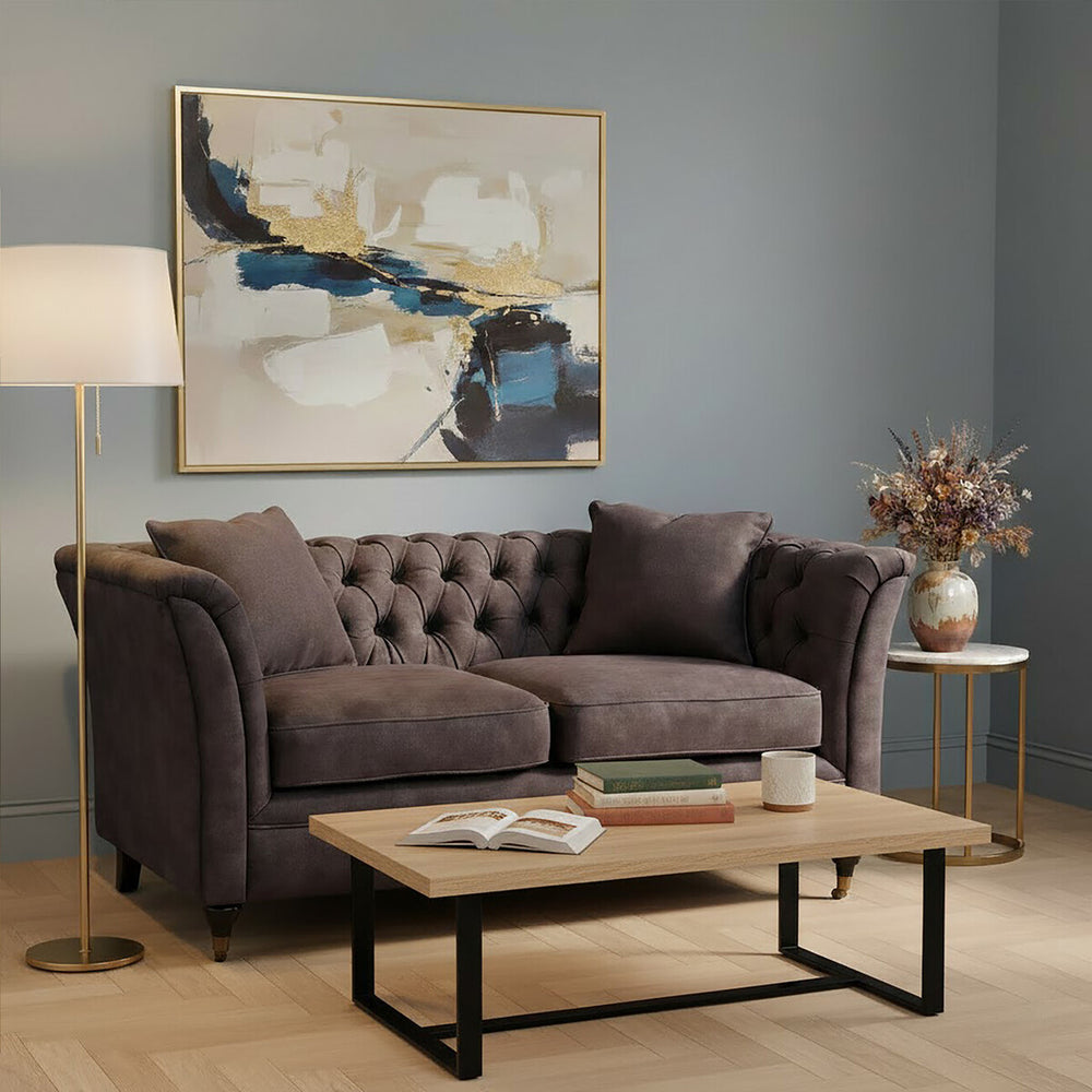 Grey velvet two-seat sofa with tufted backrest and plush cushions, positioned in a cozy living room setting. A wooden coffee table with a black metal frame sits in front of the sofa, and a floor lamp provides warm lighting. A decorative vase with flowers and an abstract wall art piece are visible in the background against a soft blue wall
