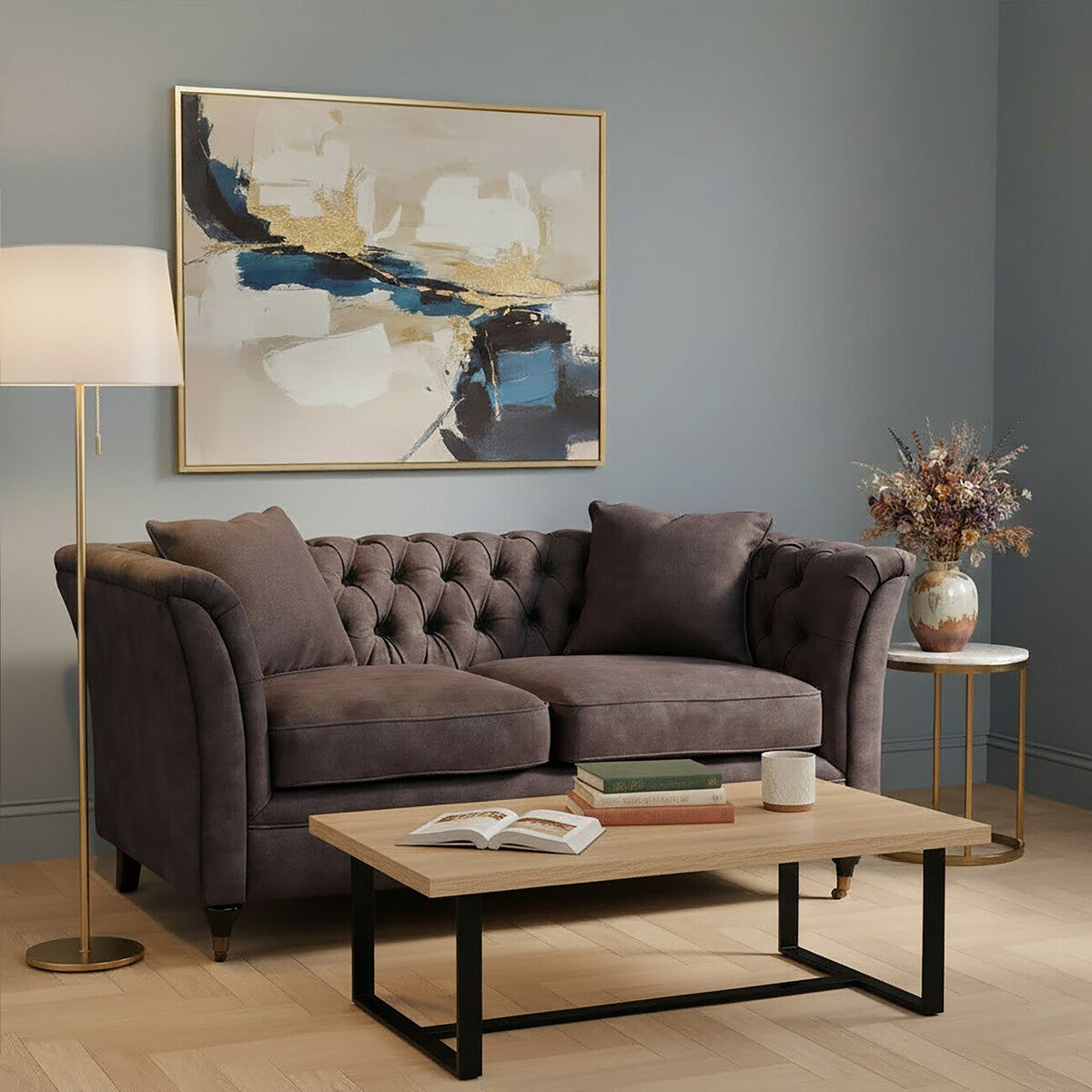 Grey velvet two-seat sofa with tufted backrest and plush cushions, positioned in a cozy living room setting. A wooden coffee table with a black metal frame sits in front of the sofa, and a floor lamp provides warm lighting. A decorative vase with flowers and an abstract wall art piece are visible in the background against a soft blue wall
