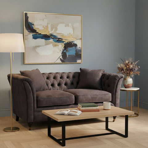 Grey velvet two-seat sofa with tufted backrest and plush cushions, positioned in a cozy living room setting. A wooden coffee table with a black metal frame sits in front of the sofa, and a floor lamp provides warm lighting. A decorative vase with flowers and an abstract wall art piece are visible in the background against a soft blue wall