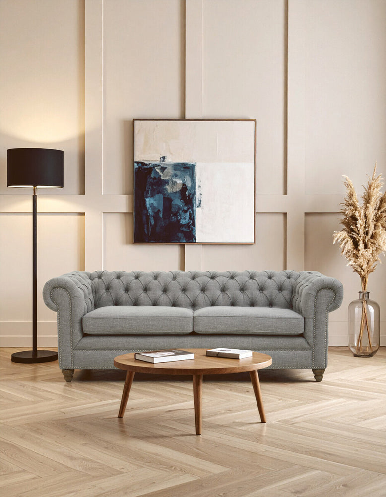 Three-seat grey linen sofa with tufted backrest, positioned in a stylish living room setting. A floor lamp stands beside the sofa, and a wooden coffee table is in front, adorned with decorative items. A large framed artwork hangs on the wall behind the sofa, complemented by a vase of dried flowers in the corner. The room features light-colored walls and a geometric patterned floor