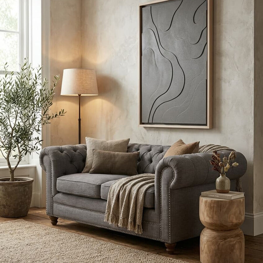 A two-seat grey linen sofa with plush cushions, adorned with decorative pillows, is positioned in a cozy living room. The sofa is complemented by a large abstract wall art piece, a floor lamp with a warm glow, and a potted plant. A textured area rug and two round wooden side tables add to the inviting atmosphere. Natural light streams in through a window, enhancing the soft color palette
