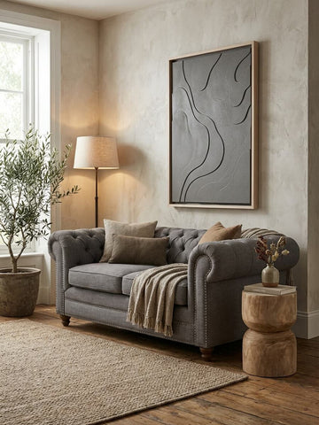 A two-seat grey linen sofa with plush cushions, adorned with decorative pillows, is positioned in a cozy living room. The sofa is complemented by a large abstract wall art piece, a floor lamp with a warm glow, and a potted plant. A textured area rug and two round wooden side tables add to the inviting atmosphere. Natural light streams in through a window, enhancing the soft color palette