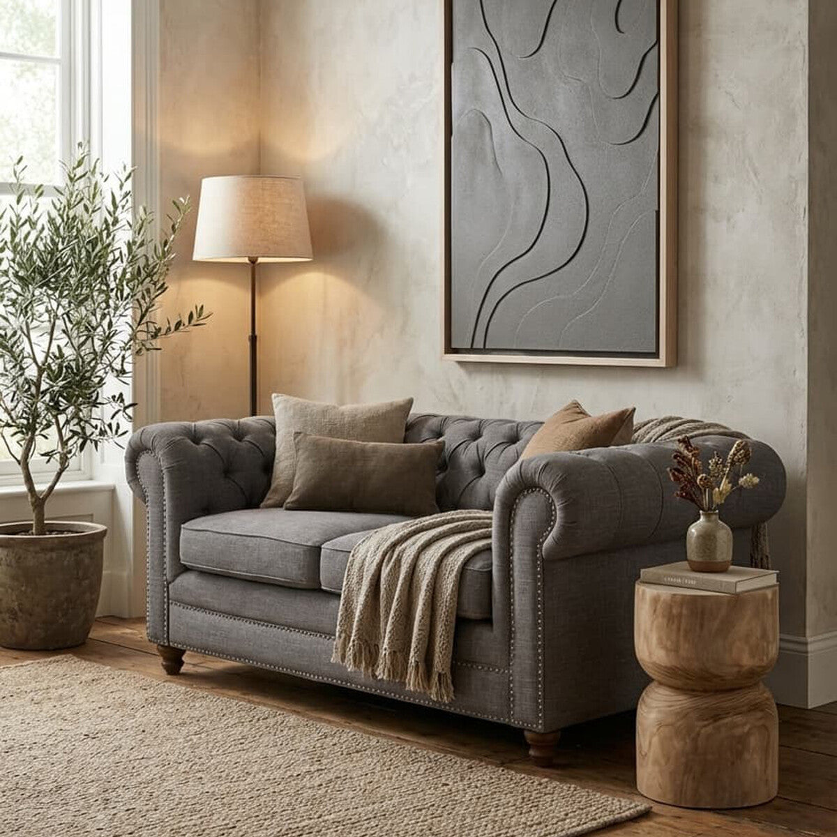 A grey linen two-seat sofa with tufted detailing, adorned with decorative pillows and a cozy throw blanket, positioned in a stylish living room setting. A tall lamp and a potted plant are nearby, with a textured area rug and wooden side tables completing the warm, inviting atmosphere