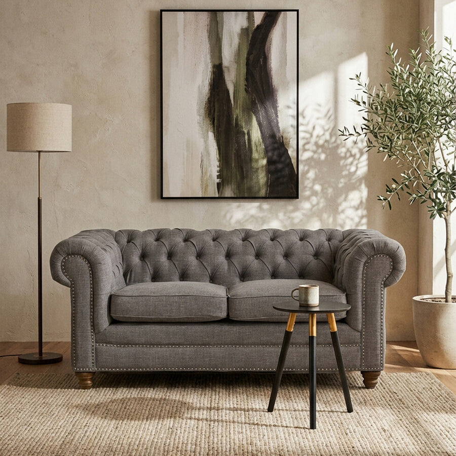 Two-seat grey linen sofa with tufted backrest, placed in a bright room featuring a large abstract wall art piece, a standing lamp, and a small round side table with decorative items, surrounded by a light-colored rug and a potted plant