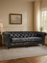 Antique black Chesterfield leather sofa with three seats, featuring tufted backrest and armrests, set against a light-colored wall with a framed artwork and a lamp on a side table, complemented by a soft area rug on the floor