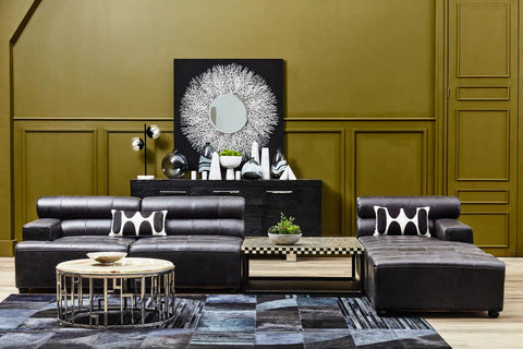 Antique black leather modular chaise sofa with a right arm, featuring a sleek design and plush cushions, positioned in a stylish living room setting. A large decorative clock and modern decor elements are visible in the background, along with a round coffee table and a patterned area rug. The walls are painted a warm gold color, enhancing the overall elegance of the space