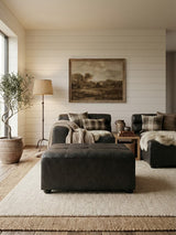 A black leather modular ottoman sits in a cozy living room setting, surrounded by a light-colored rug, a sofa with decorative pillows, and a large painting on the wall. Natural light streams in through a window, illuminating a floor lamp and a potted plant nearby