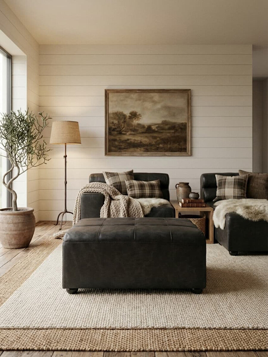 A black leather modular ottoman sits in a cozy living room setting, surrounded by a light-colored rug, a sofa with decorative pillows, and a large painting on the wall. Natural light streams in through a window, illuminating a floor lamp and a potted plant nearby