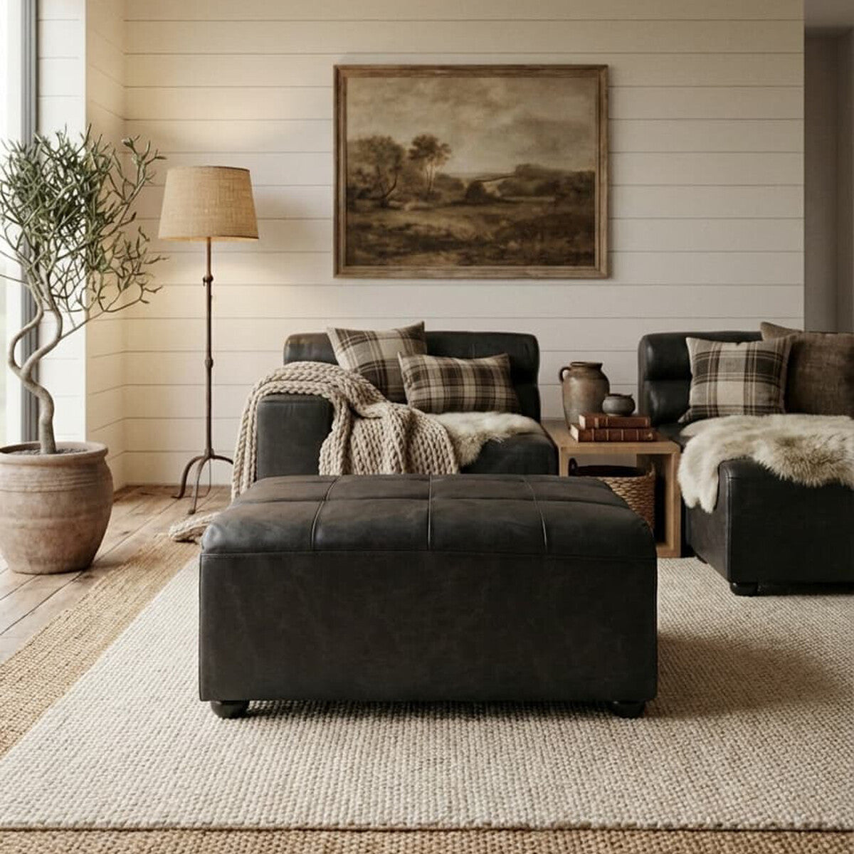 A black leather modular ottoman sits in a cozy living room, surrounded by a light-colored rug. The space features a large window with natural light, a potted plant, a floor lamp, and a sofa adorned with patterned cushions. A framed artwork hangs on the wall, adding a touch of warmth to the decor
