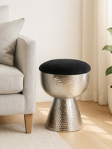A metallic stool with a sleek, shiny surface and a round black upholstered top, positioned next to a light-colored sofa in a bright, airy room with wooden flooring