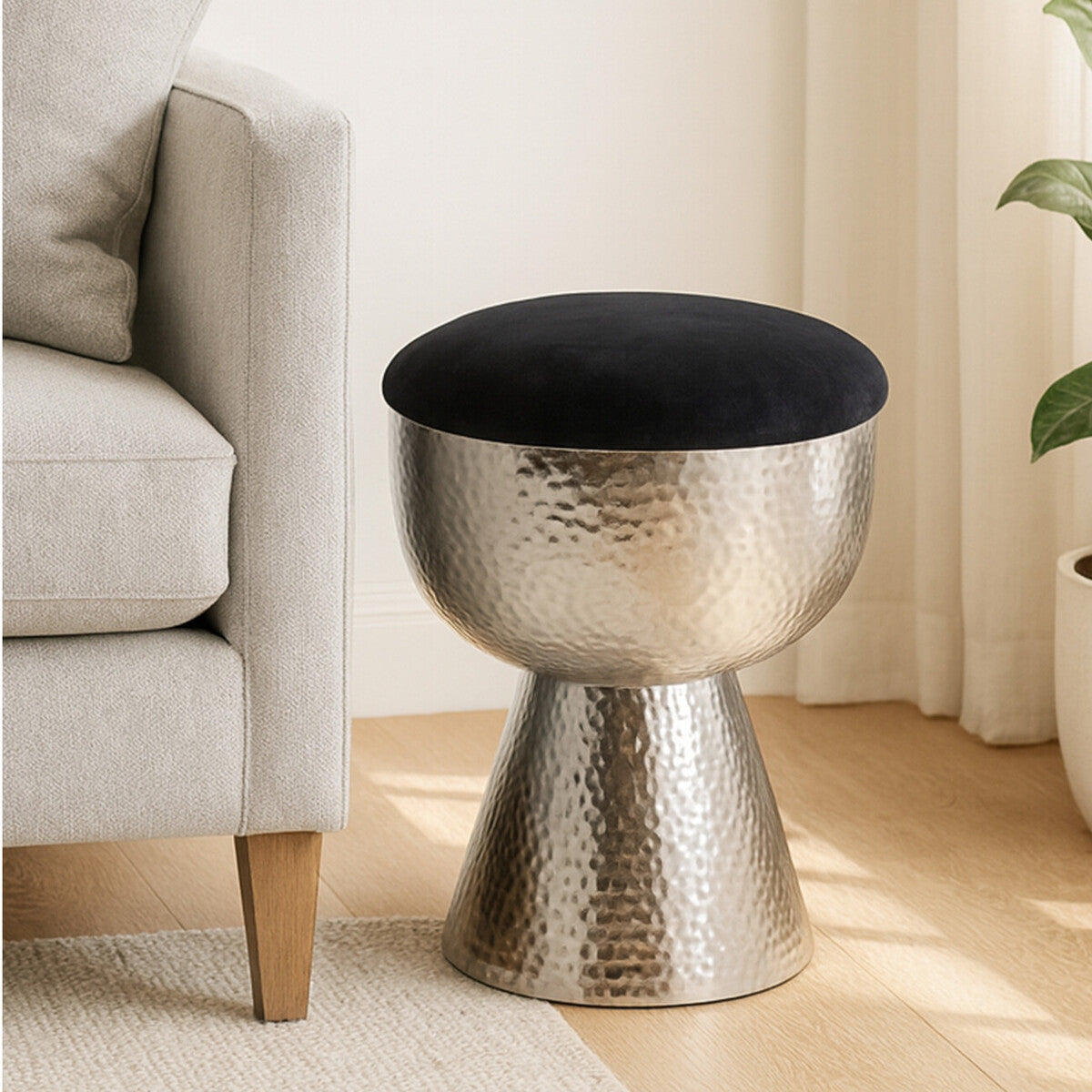 A metallic stool with a hammered finish and a round black velvet cushion on top, positioned next to a light-colored sofa in a bright room with wooden flooring