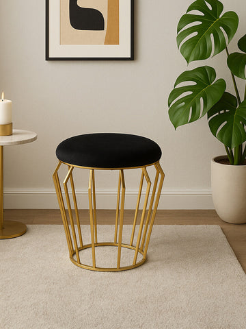 A black velvet stool with a gold finish metal base, featuring a round top and an open geometric design. The stool is placed on a light-colored rug in a modern interior setting, with a framed artwork and a potted plant visible in the background