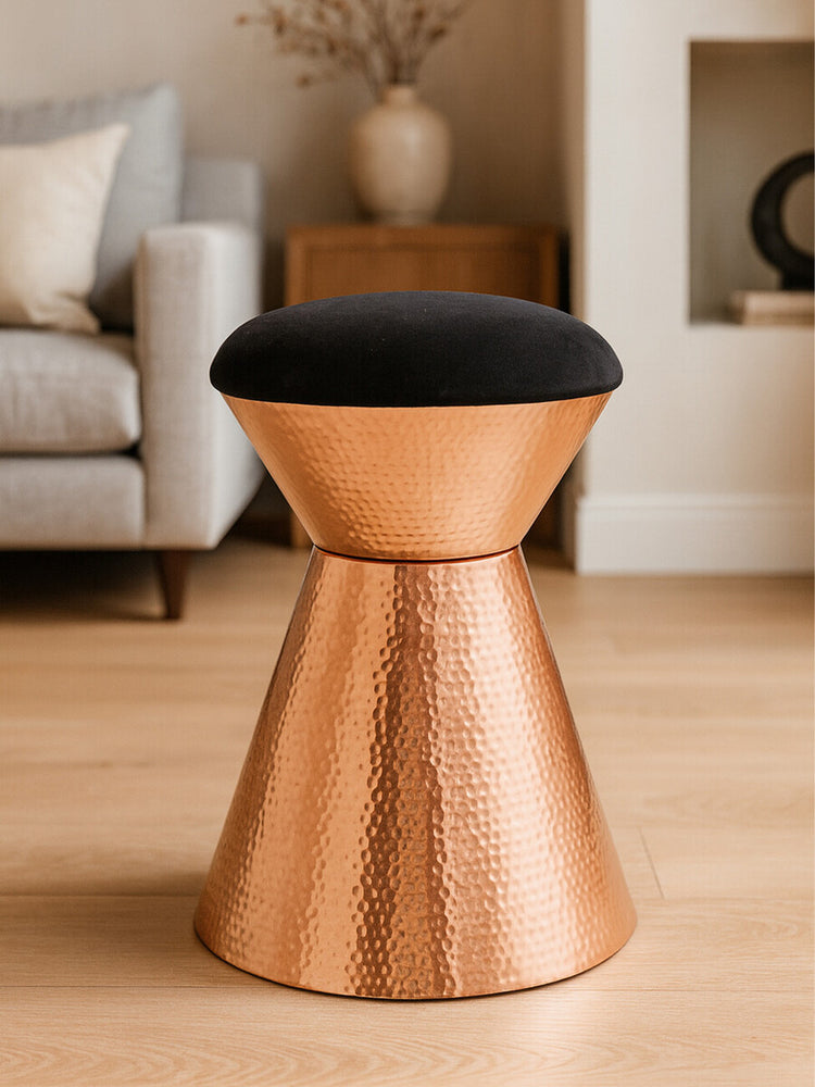 A black velvet upholstered stool with a copper finish base, featuring a modern, tapered design. The stool is placed on a light wooden floor, with a soft sofa and decorative elements visible in the background
