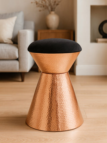 A black velvet upholstered stool with a copper finish base, featuring a modern, tapered design. The stool is placed on a light wooden floor, with a soft sofa and decorative elements visible in the background
