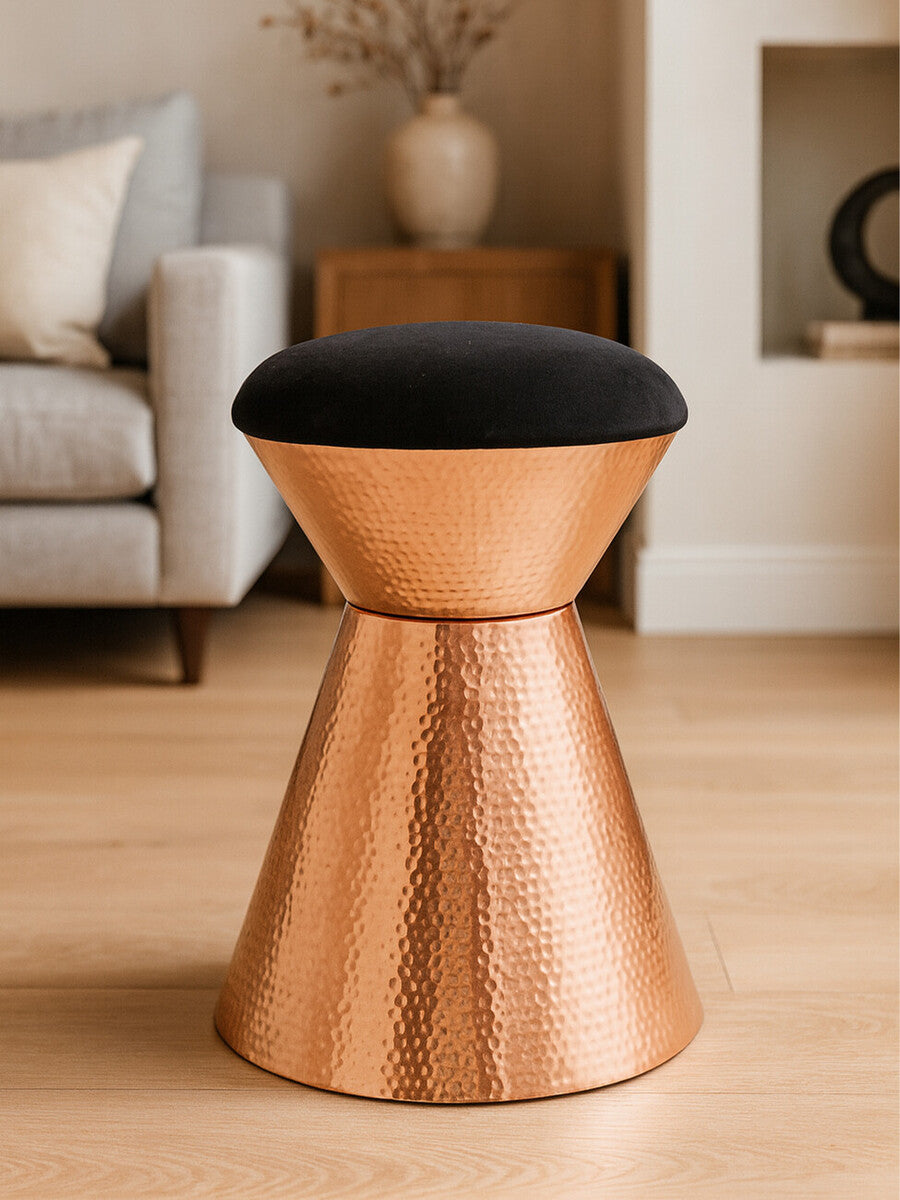A black velvet upholstered stool with a copper finish base, featuring a modern, tapered design. The stool is placed on a light wooden floor, with a soft sofa and decorative elements visible in the background