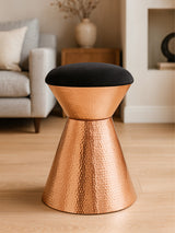 A black velvet upholstered stool with a copper finish base, featuring a modern, tapered design. The stool is placed on a light wooden floor, with a soft sofa and decorative elements visible in the background