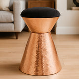 A stylish stool featuring a black velvet top and a hammered copper finish base, designed with a modern, tapered silhouette. The stool is placed on a light wooden floor, with a soft, neutral-toned sofa visible in the background