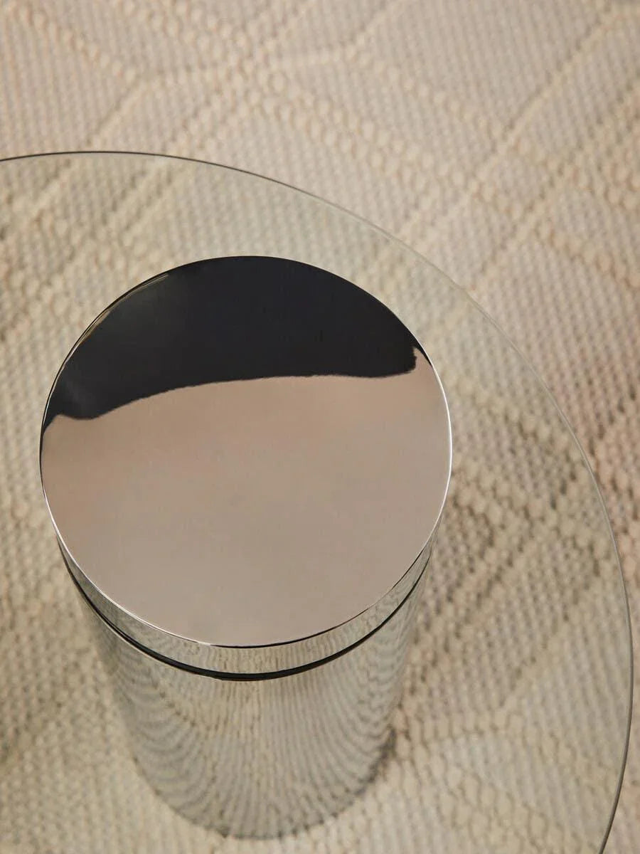 Round glass coffee table with chrome metal base on textured beige rug