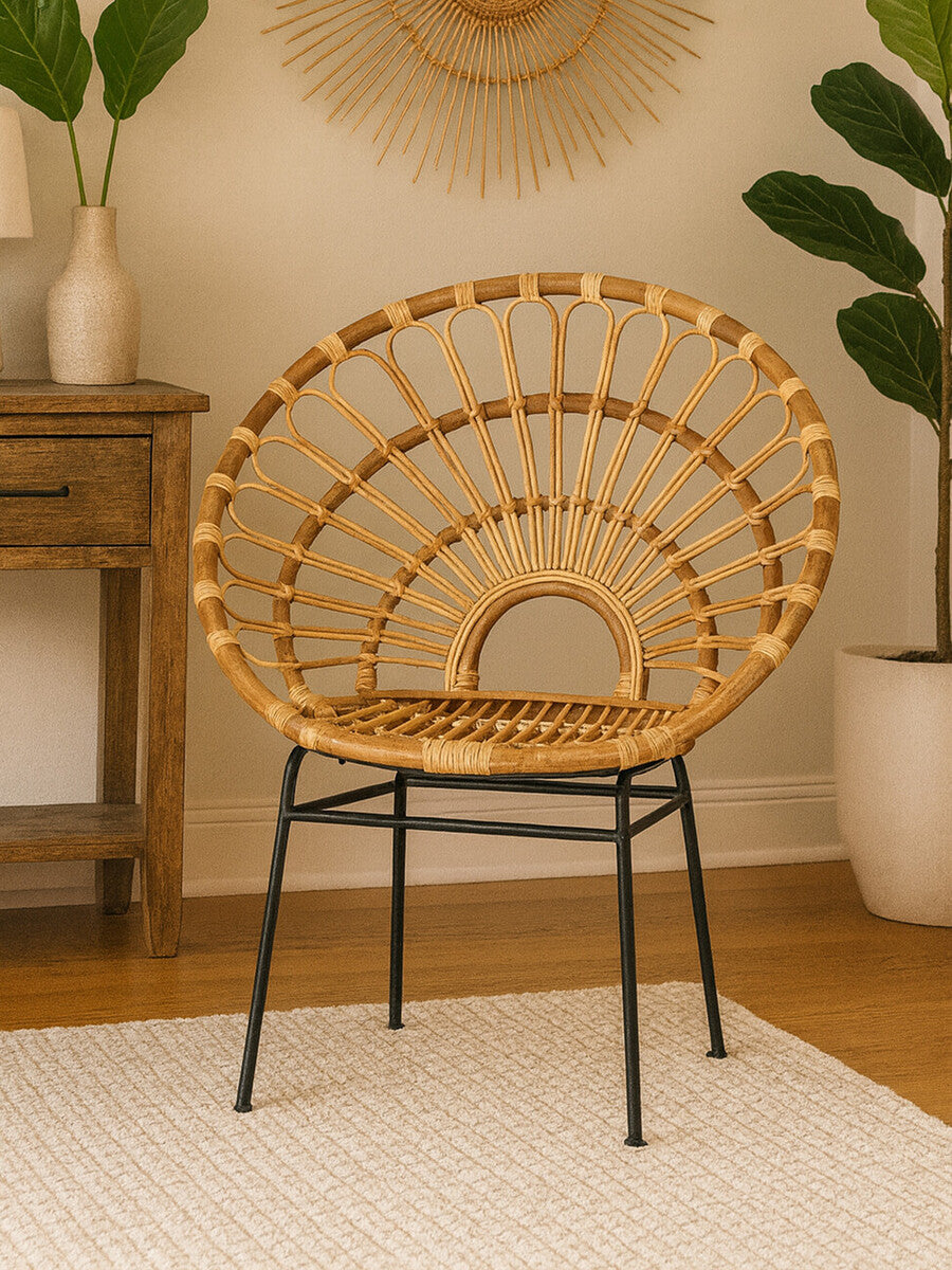 Natural rattan chair with a sunflower-inspired design, featuring a circular backrest and black metal legs, placed on a light-colored rug in a cozy interior setting with a wooden side table and potted plant in the background