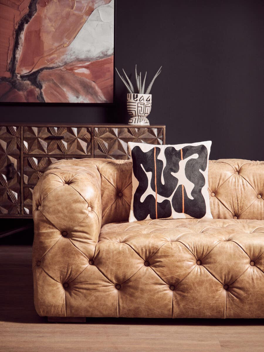 Brown leather Chesterfield sofa with tufted upholstery, featuring a single decorative black and white patterned pillow. The sofa is positioned against a dark wall, with a modern art piece and a decorative vase in the background