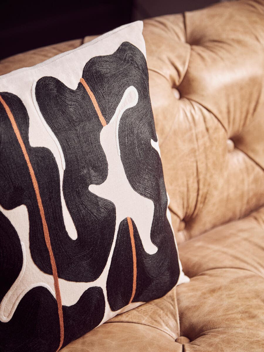 A close-up view of a decorative pillow featuring a bold black and white abstract pattern with orange accents, resting on the tufted surface of a brown leather Chesterfield sofa. The sofa's rich texture and classic design elements are highlighted in the background