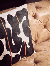 A close-up view of a decorative pillow featuring a bold black and white abstract pattern with orange accents, resting on the tufted surface of a brown leather Chesterfield sofa. The sofa's rich texture and classic design elements are highlighted in the background
