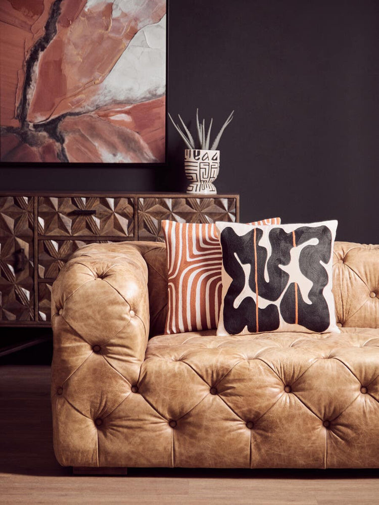Brown leather three-seat Chesterfield sofa with tufted upholstery, featuring decorative pillows in black and white patterns, set against a dark wall and a stylish abstract artwork in the background