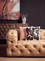 Brown leather three-seat Chesterfield sofa with tufted upholstery, featuring decorative pillows in black and white patterns, set against a dark wall and a stylish abstract artwork in the background