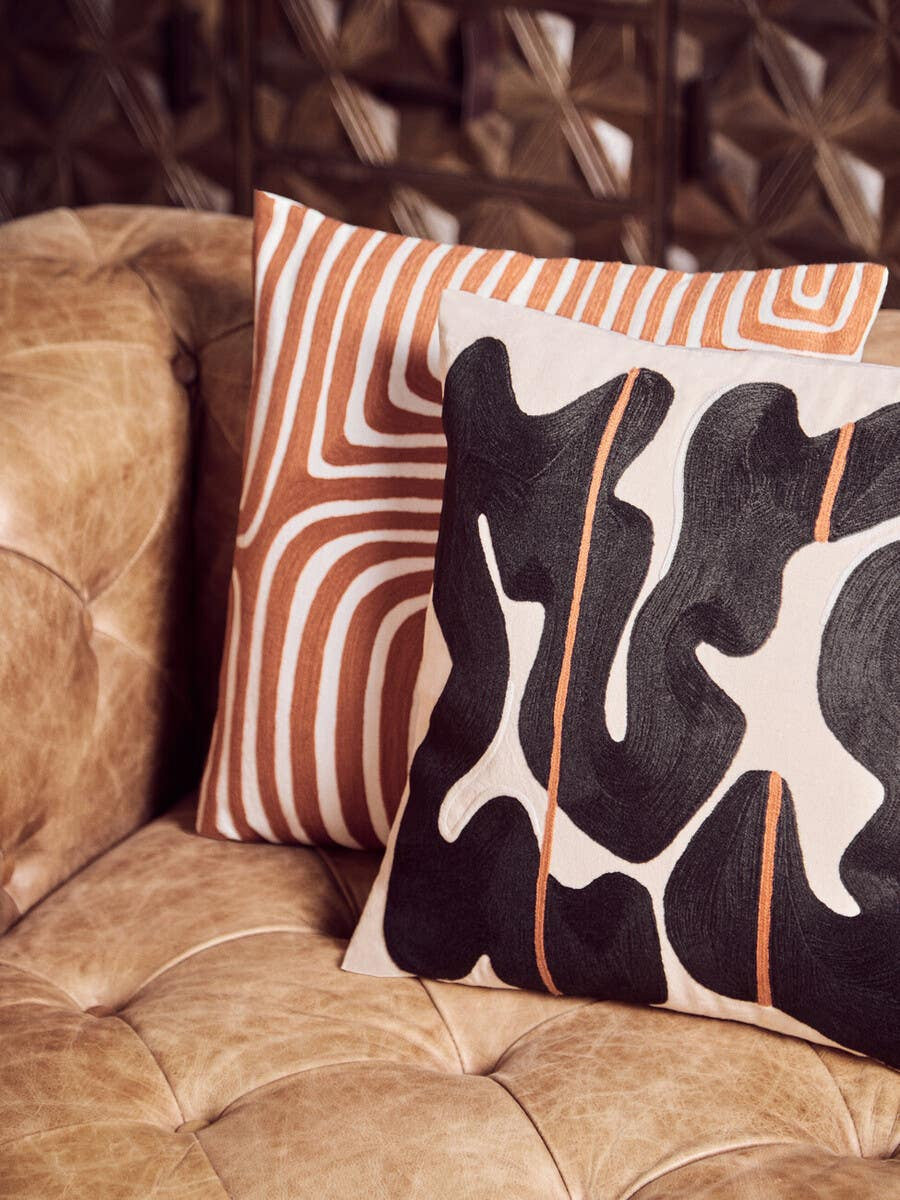 A close-up view of two decorative pillows resting on a brown leather Chesterfield sofa. One pillow features a bold black and white abstract design, while the other has a warm orange and white striped pattern. The sofa's tufted leather surface is visible in the background, showcasing its modern design