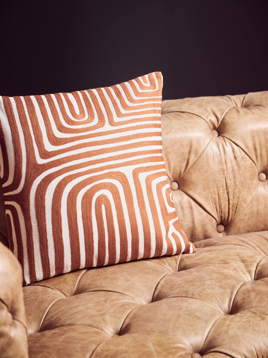 Brown leather Chesterfield sofa with tufted upholstery, featuring a decorative orange and white patterned pillow resting on the seat. The background is a dark wall, highlighting the sofa's modern design