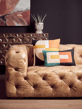 Brown leather Chesterfield sofa with tufted upholstery, featuring three seats and decorative pillows in various colors placed on one side. A modern art piece and a potted plant are visible in the background, enhancing the stylish interior setting
