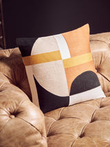 A close-up view of a decorative pillow featuring a geometric design in orange, white, and black, resting on the tufted brown leather surface of a modern Chesterfield sofa. The background is dark, highlighting the texture and color of the leather and the pillow