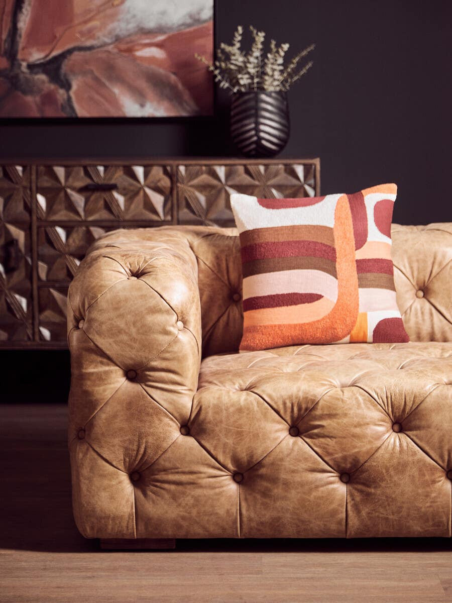 Brown leather Chesterfield sofa with a modern design, featuring tufted upholstery and a low back. Decorative pillows in warm colors are placed on the sofa, and a textured wall with a geometric pattern is visible in the background, along with a vase containing dried plants