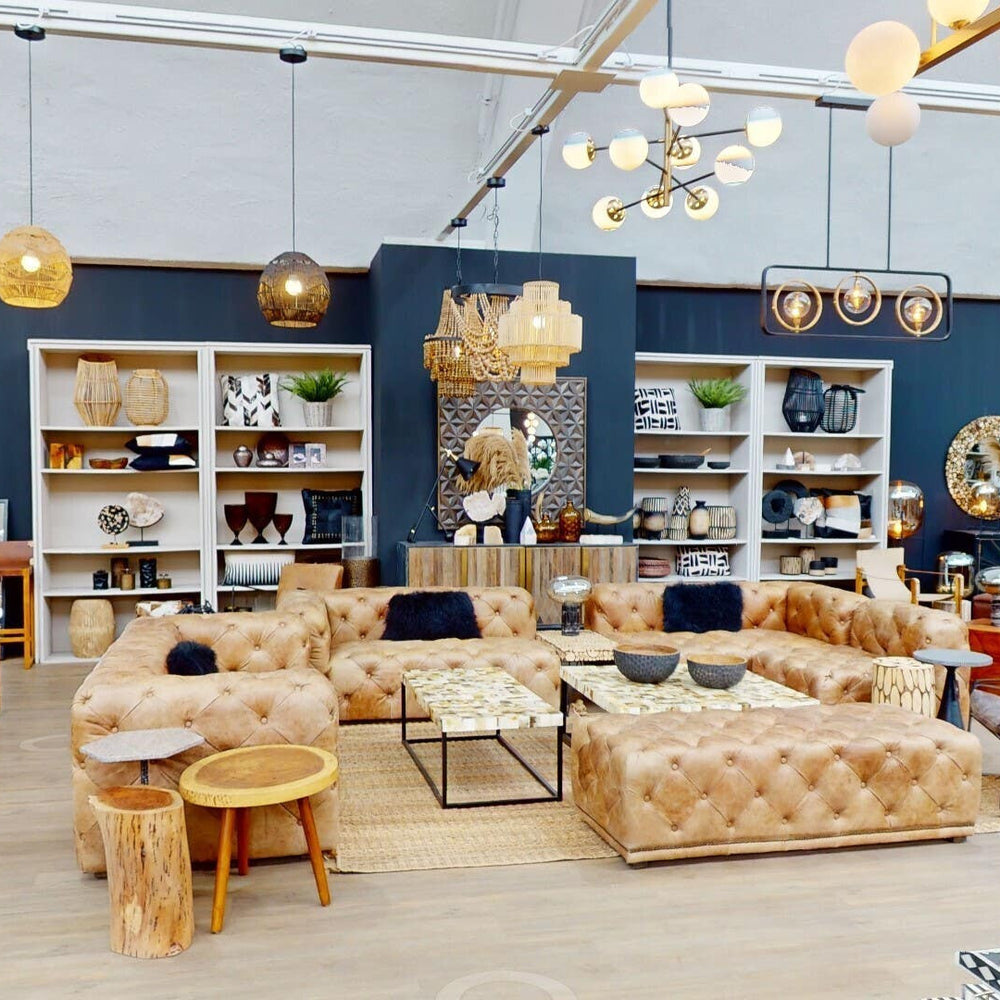 A stylish three-seat brown leather Chesterfield sofa with a modern design, featuring deep button tufting and rolled arms, placed in a bright, contemporary living space. The background includes shelves filled with decorative items and plants, along with a warm color palette and ambient lighting