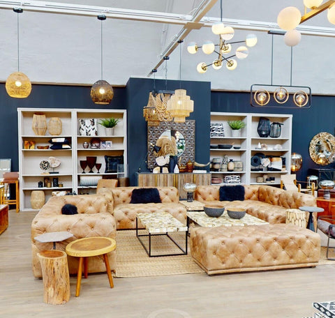 A stylish three-seat brown leather Chesterfield sofa with a modern design, featuring deep button tufting and rolled arms, placed in a bright, contemporary living space. The background includes shelves filled with decorative items and plants, along with a warm color palette and ambient lighting