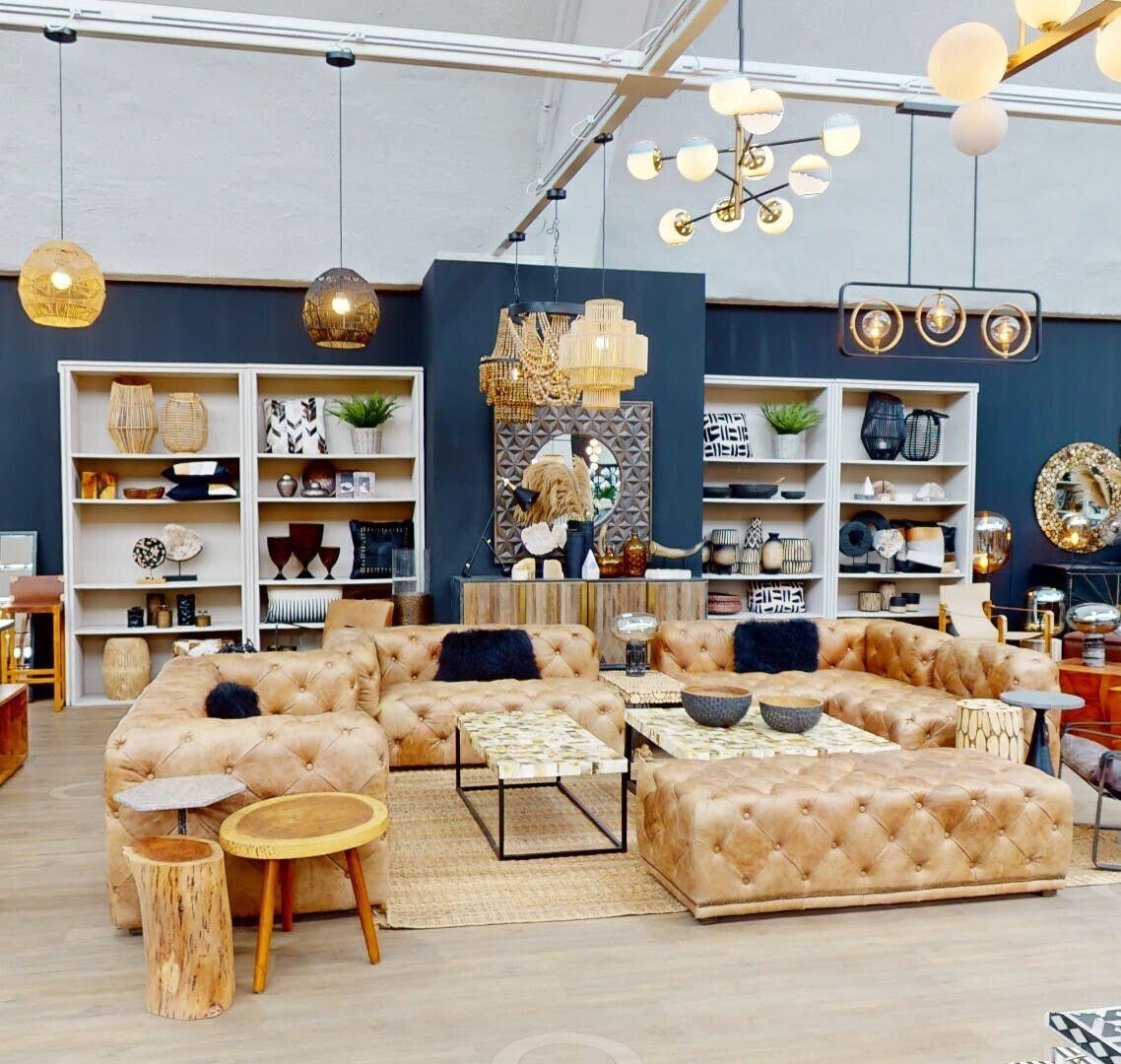 A stylish three-seat brown leather Chesterfield sofa with a modern design, featuring deep button tufting and rolled arms, placed in a bright, contemporary living space. The background includes shelves filled with decorative items and plants, along with a warm color palette and ambient lighting
